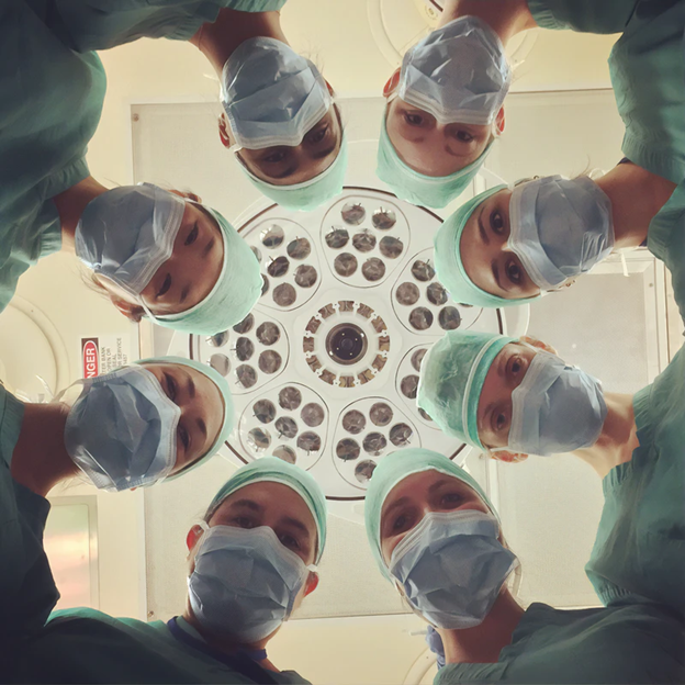 Picture of eight surgeons in light-green face masks, surgical caps, and scrubs, viewed from a patient's vantage point. They are in a circular formation, and only their heads and upper torsos can be seen.