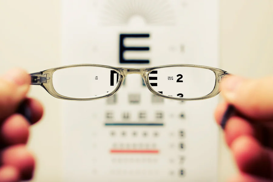 Picture of a small, grey pair of glasses being held by someone’s fingertips. Through the glasses, you can see letters and numbers on an eye chart. The rest of the eye chart is obscured.