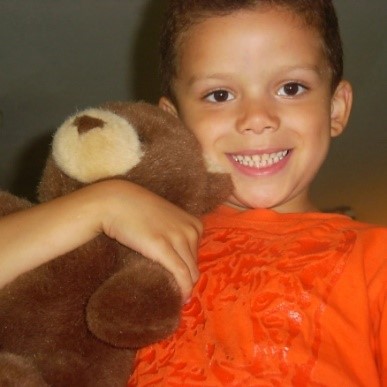 This is a picture of Miss Ruth’s son, Coleman, at age 5. Coleman has short, wavy brown hair, brown eyes, tan skin, and a joyful smile on his face. He is wearing a bright orange shirt with an applique of a tiger’s face on it. He is holding his brown teddy bear, Aaliyah. PS: Coleman is now 23, while, paradoxically, Miss Ruth is still 29 years old (although her license claims she's really 56 😉)