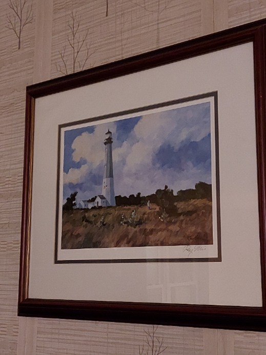 The picture is a painting of a lighthouse. There is a small white house or building to the left of the lighthouse. It is set against a blue sky with some clouds. The lighthouse appears to be on a small hill or cliff with some greenery around it. The painting is framed with dark wood and has a white mat border. The wall on which it is hung has a textured wallpaper with a subtle tree design.